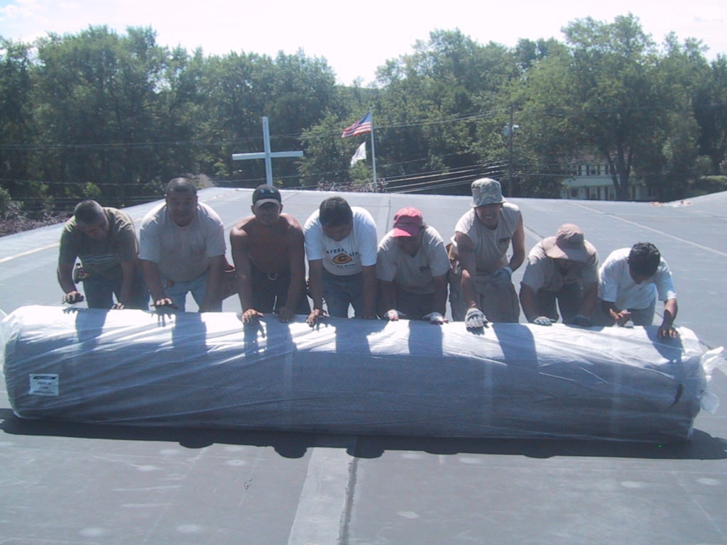 Commercial roof replacement costs can be affected by the materials chosen. Workers roll out roofing material on a roof.